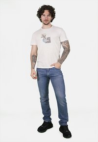 T-shirt in cotone beige chiaro con stampa grafica, abbinata a jeans in denim blu. Il modello indossa sneaker nere e ha tatuaggi visibili sulle braccia.