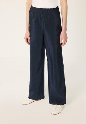 Personne portant un pantalon large bleu marine avec une ceinture élastique et des chaussures blanches décontractées à lacets, debout sur un fond uni.