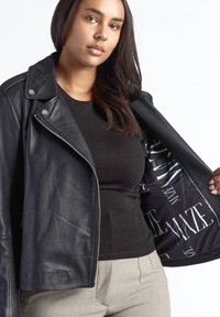 Sort læder bikerjakke med sølvfarvede metaldele og lynlåse. Indersiden har et mønstret sort foer med hvid tekst. Glat tekstur.