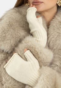 Rękawiczki bez palców z kremowego, dzianego materiału o prążkowanej fakturze; noszone na miękkim, jasnobrązowym futrze. Ręce spoczywają blisko twarzy.