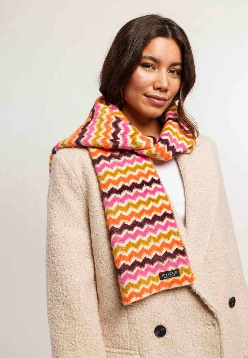 Donna che indossa un cappotto beige con texture e una sciarpa a zigzag colorata nei toni di rosa, arancione, senape e marrone, su uno sfondo semplice.