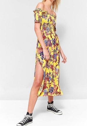 Robe jaune à fleurs avec épaules dénudées, corps smocké et fente latérale, ornée d'accents violets, rouges et verts ; portée avec des baskets noires montantes.