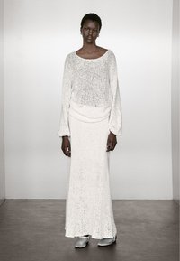 Długa biała dzianinowa sukienka o luźnym fasonie, z długimi rękawami i okrągłym dekoltem. Gładka faktura i prosty design. Dopełnieniem stroju są srebrne buty.