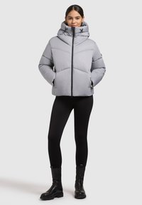Giubbotto imbottito grigio chiaro con colletto alto, chiusura con zip e dettagli di trapuntatura diagonale. Il modello indossa leggings neri e robusti stivali neri.