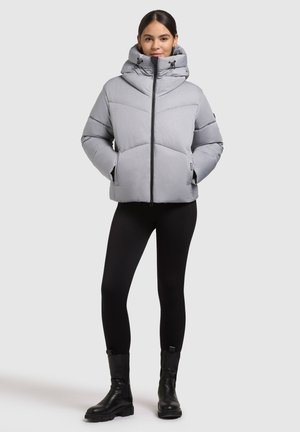 Šviesiai pilka pūkinė striukė su aukštu apykakle, užtrauktuko uždarymu ir įstrižais siuvinėjimais. Modelis dėvi juodas tampres ir tvirtus juodus batus.