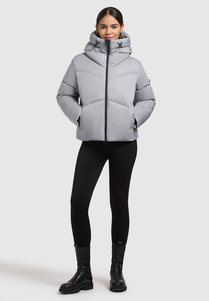 Svetlo siva pufasta jakna z visokim ovratnikom, zadrgo in diagonalnim šivom. Model nosi črne legice in trdne črne škornje.