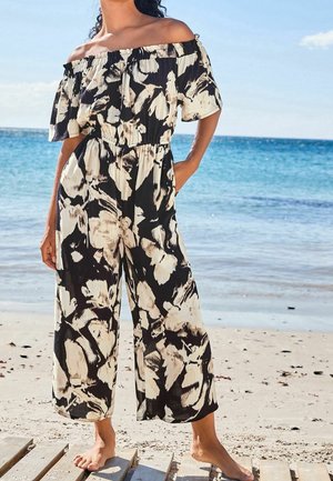 Sort og cremefarvet buksedragt med blomsterprint, off-shoulder design, vide ben og rynket talje, sat mod en strandbaggrund.