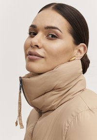 Beige pufferjacka med hög krage, som har dragkedja och diskret horisontell sömnad. Modellen har guldörhängen och slät hy.