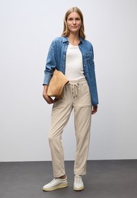 Frau mit einem weißen Oberteil, einer Jeansjacke, beigen Jogginghosen, weißen Sneakers, die eine tanfarbene Wildleder-Clutch vor einem schlichten Hintergrund hält.