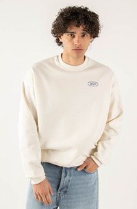 Felpa beige chiaro con colletto rotondo e polsini a coste. Presenta un piccolo logo blu ovale sul petto. Indossata con jeans in denim blu.