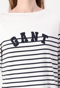 Balta ir tamsiai mėlyna stripes marškiniai su laivo kaklu, kuriuose krūtinėje yra išsiuvinėtas žodis "GANT" tamsiai mėlynomis siūlėmis. Minkšta tekstūra.