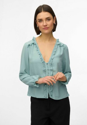 Žena s krátkymi tmavými vlasmi oblečená v svetlomodrej blúzke s volánikmi a čiernych nohaviciach, stojaca s jemne zloženými rukami.
