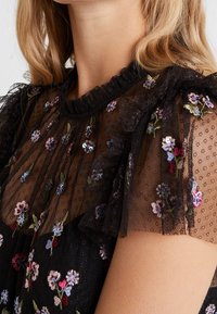 Blouse noire avec broderies florales, en tissu transparent à pois, avec un col et des manches volants, présentant des motifs de fleurs multicolores et des sequins.