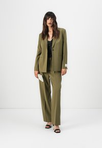 Olive green tailored suit with a single-button blazer and wide-leg trousers. Worn over a black top, paired with a textured white clutch.
