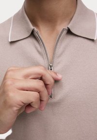 Beige polo-shirt med lynlås og tekstureret stof, med en kontrasterende hvid krave og en glat lynlås, delvist åben med et greb fra hånden.