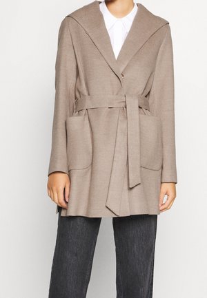 Persoon draagt een beige jas met riem en grote voorkantzakken over een wit overhemd en zwarte broek tegen een effen achtergrond.