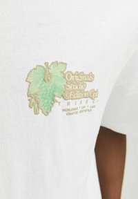 Balta medvilnė marškinėliai su žaliu ir smėlio spalvos siuvinėtą vynuogių dizainą, kuriame yra tekstas "Originals Studio Edition" ir "Tribeca".