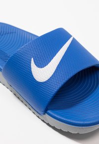 Blå sklisandal med teksturert overdel, med en hvit Nike swoosh. Grå såle med ribbet tekstur for grep.