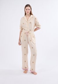 Lichtbeige gestreepte pyjamaset met bloemenborduursel. Bevat een knoopsluiting shirt, elastische taillebroek en roze manchet details.