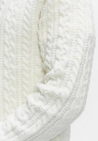 Valkoinen neulottu kangas, jossa on yksityiskohtaiset kaapelikuviot ja resorit, pehmeän ja teksturoidun neulepuseron hiha.