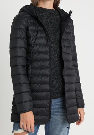Femme portant une veste matelassée noire à capuche par-dessus un pull gris foncé et un jean bleu déchiré sur un fond uni.