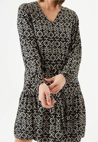 Svart och crème mönstrad klänning med V-ringning, långa ärmar, samlade manschetter och en lager-på-lager kjoldesign. Slät tyg med geometriska motiv.