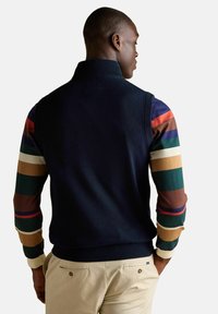 Mężczyzna w granatowej kamizelce bez rękawów na tle wielokolorowej, paskowanej koszuli z długim rękawem i beżowych spodniach, widziany z tyłu.