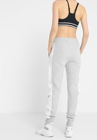 Graue Jogginghose mit weißen Seitenstreifen, elastischem Bund und gerippten Bündchen. Kombiniert mit einem schwarzen Sport-BH und weißen Turnschuhen.