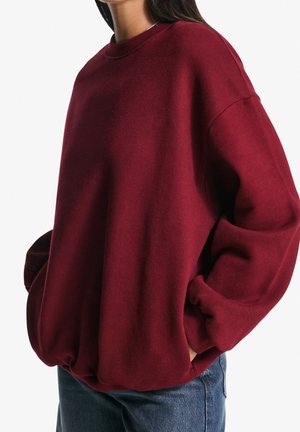 Femme portant un pull bordeaux ample à manches longues, les mains dans les poches, associé à un jean bleu en denim.