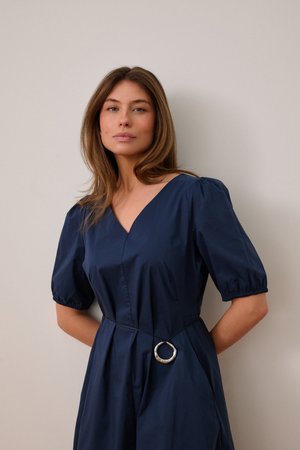 Vrouw met steil lichtbruin haar, gekleed in een marineblauwe jurk met pofmouwen en een zilveren ronde riemdetail, staand tegen een effen achtergrond.