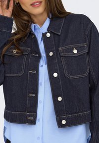 Femme portant une veste en jean foncé avec des boutons argentés sur une chemise bleu clair boutonnée, touchant ses cheveux près de l'oreille avec une main.