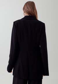 Černý blazer s přiléhavým střihem, strukturovanými rameny a dlouhými rukávy. Hladká tkanina s čistým, minimalistickým designem.