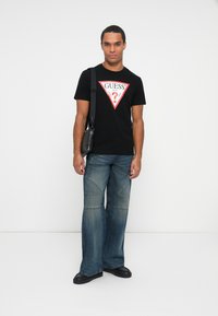 Tricou din bumbac negru cu logo-ul "GUESS" roșu și alb, asociat cu blugi deschiși la culoare, cu picior larg, și încălțăminte neagră.