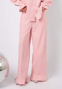 Stripete rosa og hvite bukser med vide ben og volangkant. Lett stoff, løs passform og subtil horisontal tekstur. Sølvfargede sko i nærheten.