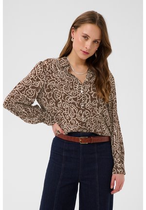 Jeune femme portant un chemisier marron à manches longues à motif cachemire, rentré dans un jean bleu foncé taille haute avec une ceinture en cuir marron, posant avec la main sur la hanche.