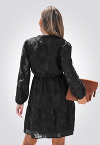 Vestido negro con estampado floral, de manga larga, cintura elástica y falda con volantes. La modelo sostiene un bolso de mano texturizado marrón.