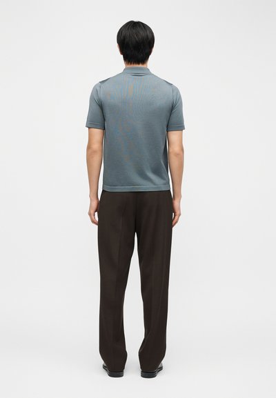 Άνδρας που στέκεται με την πλάτη, φορώντας μπλε πόλο με κοντό μανίκι και φαρδιά μαύρα παντελόνια, σε λευκό φόντο.