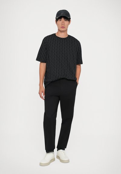 T-shirt graphique noir avec un motif répétitif, associé à un pantalon noir et des baskets blanches avec une semelle grise. Vêtu de manière décontractée.