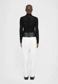 Black sheer top with repeated print and long sleeves, paired with white vertically striped wide-legged trousers and chunky black shoes.