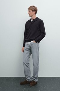 Young man with short hair stands against plain wall wearing black long-sleeve polo, light gray jeans, brown shoes, one hand in pocket.