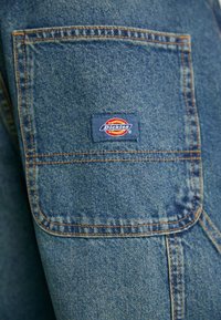 Modra denim tkanina z vidnimi šivi. Na voljo je žep s pravokotno oznako Dickies v rdeči, rumeni in modri barvi.