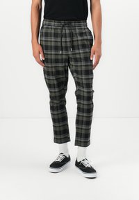 Kariert Jogginghosen in Schwarz, Grau und Olivgrün. Mit einem elastischen Bund und Kordelzug sowie schmal zulaufenden Beinen. Getragen mit schwarzen Turnschuhen.