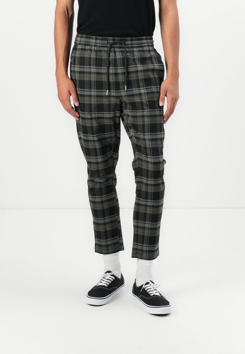 Kariert Jogginghosen in Schwarz, Grau und Olivgrün. Mit einem elastischen Bund und Kordelzug sowie schmal zulaufenden Beinen. Getragen mit schwarzen Turnschuhen.