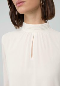Woman wearing a high-neck cream blouse with a small vertical slit and pintuck details, paired with medium hoop earrings.
