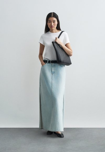 Gonna maxi in denim azzurro chiaro con spacchi laterali, abbinata a una maglietta bianca aderente e cintura nera. Borsa a spalla nera in tessuto strutturato e scarpe basse.
