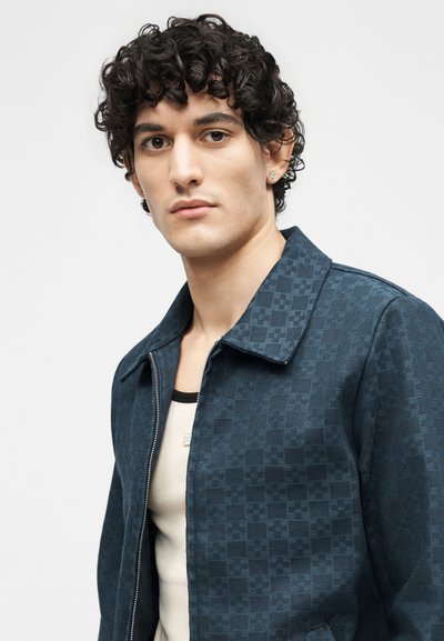 Νέος άνδρας με σγουρά σκούρα μαλλιά, φορώντας μπλε σκούρο καρό μπουφάν με φερμουάρ πάνω από ένα ανοιχτόχρωμο πουκάμισο, κοιτάζοντας μπροστά με ουδέτερη έκφραση.