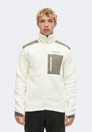 Junger Mann mit kurzen blonden lockigen Haaren, der eine weiße Fleecejacke mit grauen Schulterpatches und einer Reißverschlusstasche auf der Brust trägt.