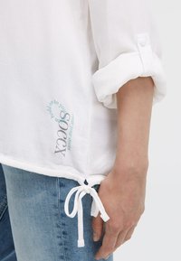 Weißes Langarmhemd mit gerollten Manschetten, Knoten-Detail am Saum und kreisförmigem Textdesign in der Nähe der Taille, kombiniert mit hellblauen Jeans.