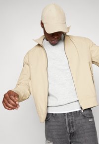 Chaqueta bomber beige con cierre de cremallera, llevada sobre un sudadera gris claro. Jeans negros desgastados y una gorra beige a juego completan el conjunto.