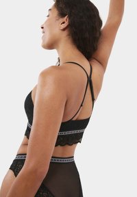 Bralette en dentelle noire avec un ourlet festonné, des bretelles ajustables et une bande contrastante arborant un motif logo. Texture lisse et douce.